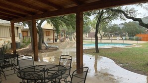 A covered patio area with metal tables and chairs, a concrete floor, and a view of a swimming pool and surrounding landscape. - Double L River House @ Frio Premier Rental Homes (Concan)