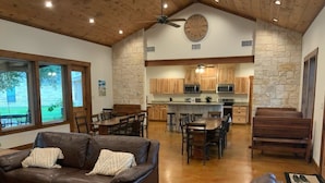 A spacious living area with a brown leather sofa, a dining table with chairs, and a kitchen area with wooden cabinets and a stone wall. - Double L River House @ Frio Premier Rental Homes (Concan)