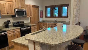 A kitchen with granite countertops, a built-in microwave, a double oven, wooden cabinets, a stone accent wall, a window with a view, and a bar area with stools. - Double L River House @ Frio Premier Rental Homes (Concan)