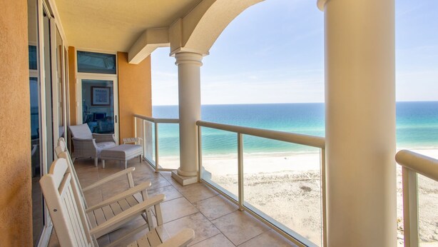 Un balcón con vista a la playa, columnas blancas y un espacio de estar con sillas y una mesa.