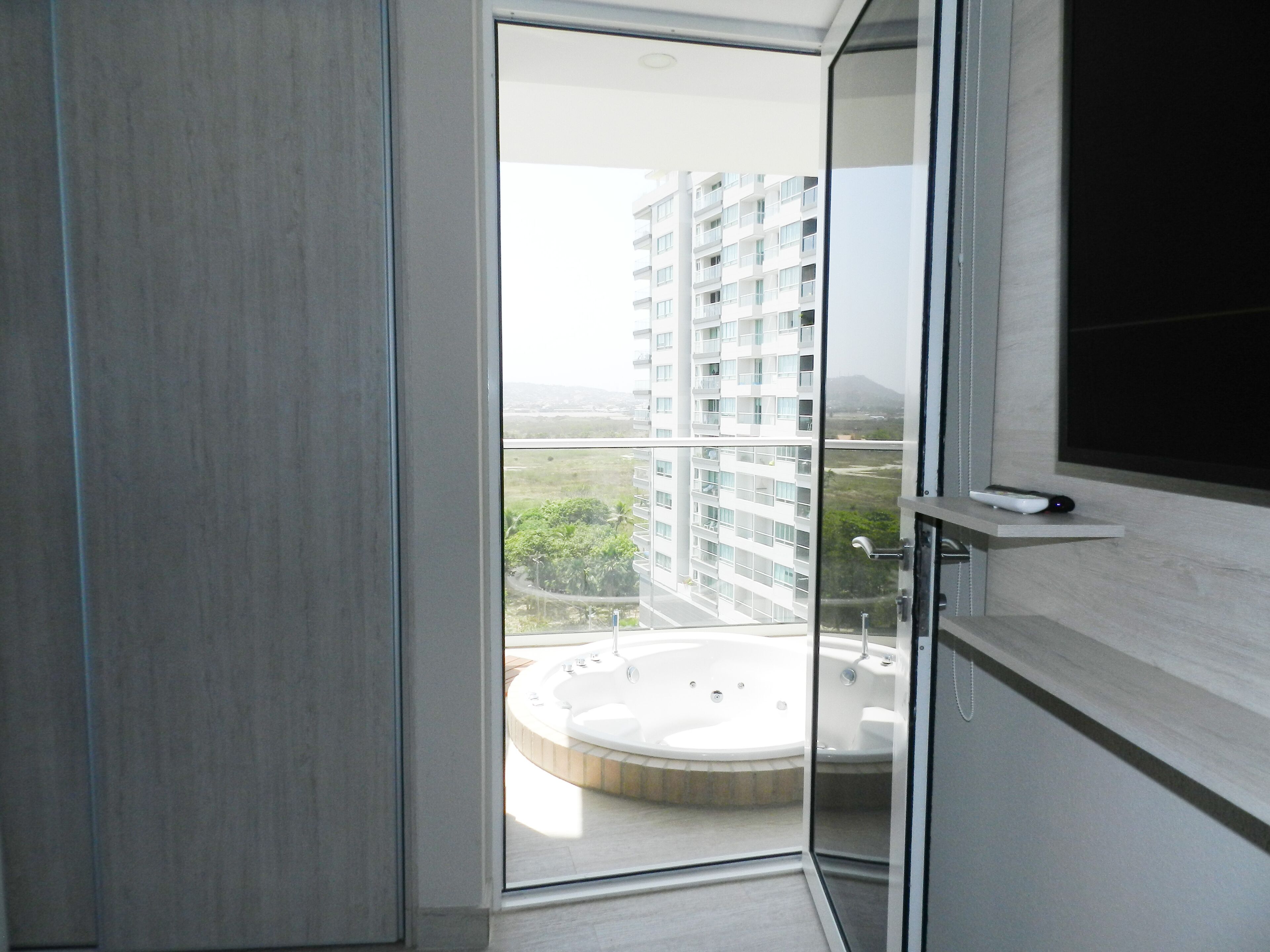 Un baño moderno con puerta de cristal que da a un balcón. El balcón ofrece vistas a un edificio alto y una bañera de hidromasaje circular.
