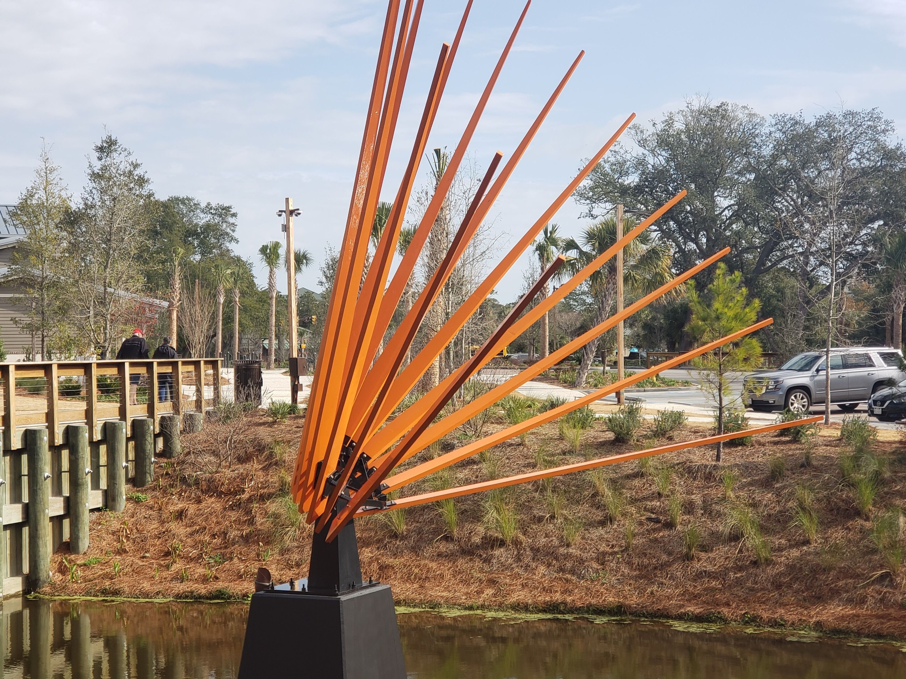 Una scultura di bastoncini arancioni in un parco con uno stagno, alberi e un ponte di legno.