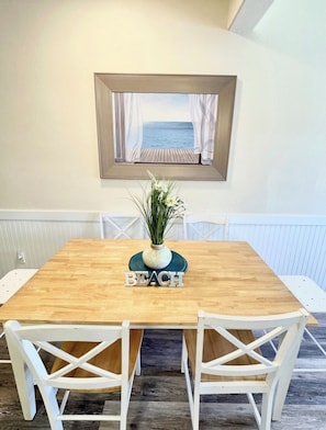 Une table à manger ornée d’une pancarte indiquant « PLAGE », de chaises blanches et, au mur, d’un tableau représentant un paysage de plage.