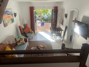 Un salon comprenant un canapé, une table basse et un téléviseur. Il y a également un coin repas avec une table et des chaises. La pièce est dotée d’une grande fenêtre et d’une porte donnant sur l’extérieur.