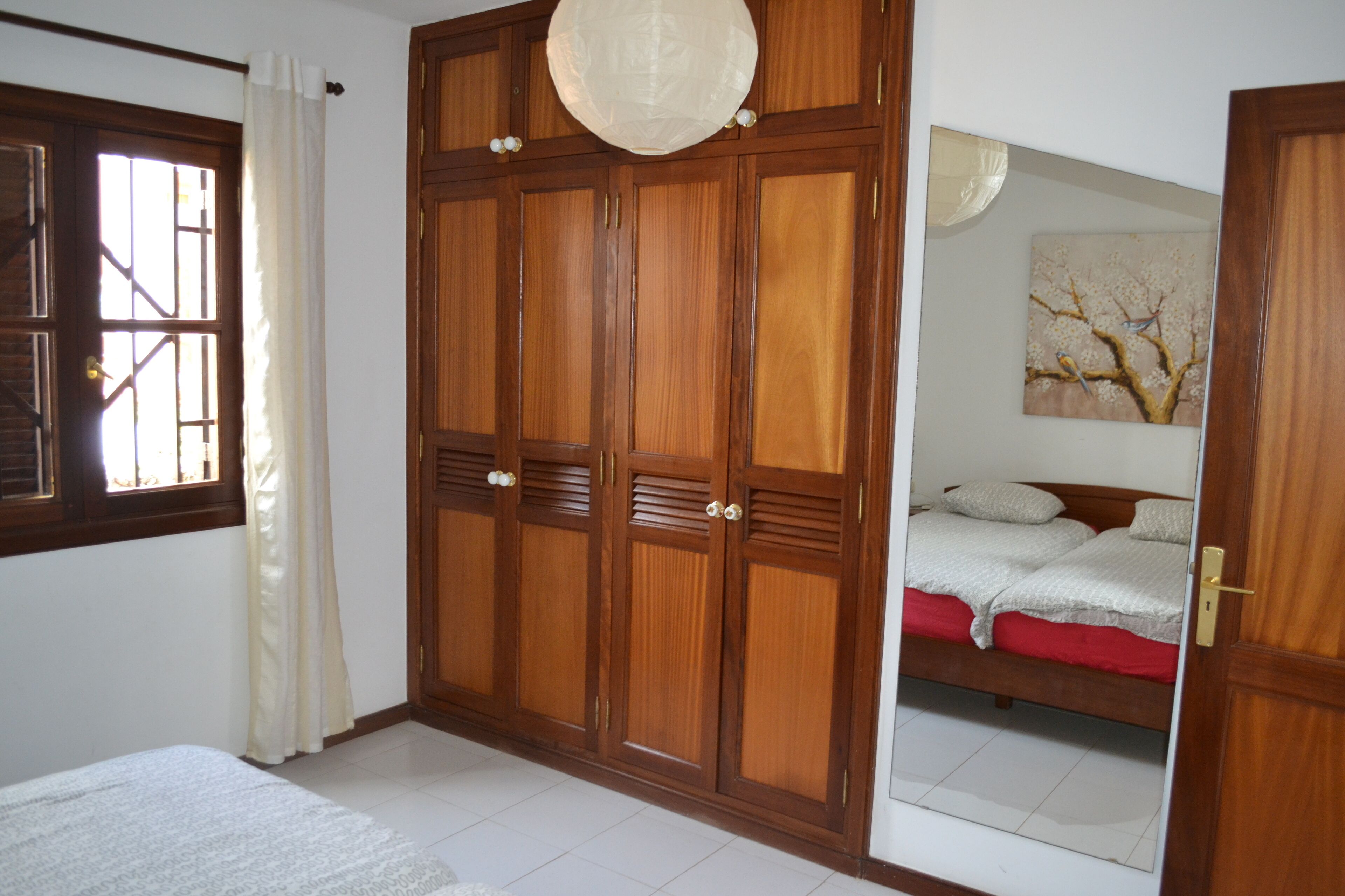 A bedroom with a wooden wardrobe, a bed with red bedding, and a painting on the wall.