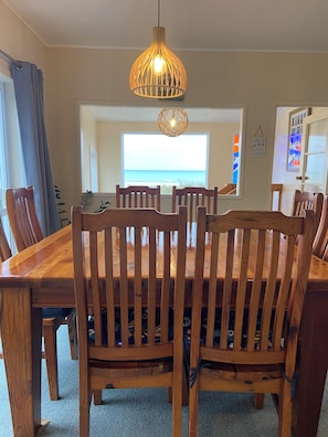 A dining area with a wooden table and chairs, a hanging light fixture, a view of the ocean through a window, and a wall-mounted clock.
