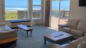 A spacious living room with a large sliding glass door leading to a balcony with a view of the ocean. There are two sofas, a coffee table, and a small table with chairs.