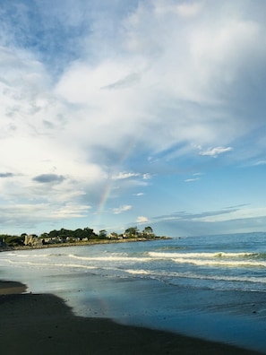 A beach with waves, a rainbow, and a row of houses in the background.