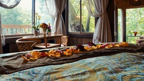 Ein Schlafzimmer mit einem Bett, einem Fenster mit Vorhängen, einem Holzstuhl, einem kleinen Tisch mit Blumen und Blick auf Bäume.