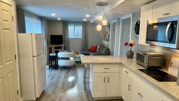 A modern kitchen with white cabinetry, a built-in microwave, a toaster, and a stove. There is a dining area with a table and chairs, and a living room with a sofa and a TV.