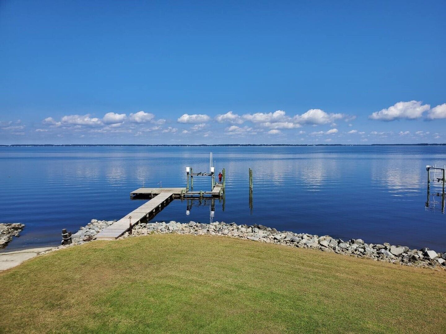 A calm lake with a wooden dock extending into the water, surrounded by a grassy area and a rocky shoreline.