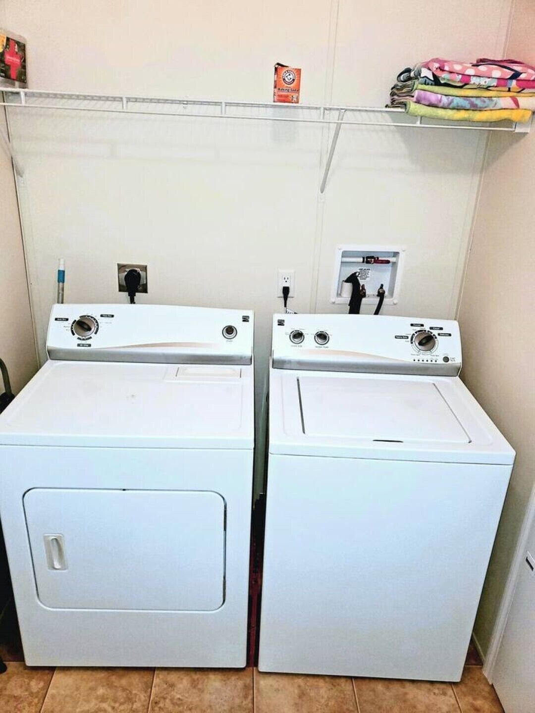 Deux machines à laver à chargement frontal, de couleur blanche, se trouvent dans une buanderie. Au-dessus, une étagère contient des vêtements pliés et une petite boîte.