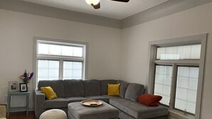 Uma sala de estar moderna com um sofá seccional cinza, dois pufes, uma mesinha de centro com um vaso e um ventilador de teto.