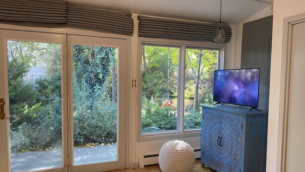Um quarto com uma janela grande, uma televisão sobre um móvel e um banquinho redondo.
