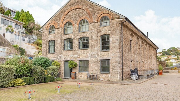 A stone building with a steeply pitched roof, multiple windows, and a small garden area with a bench and potted plants.