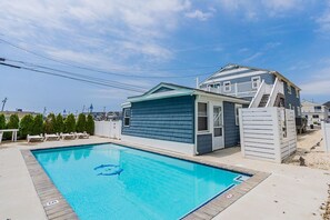 Une piscine avec une terrasse bleue, un petit bâtiment doté d’un toit blanc et un escalier menant à une maison à l’extérieur bleue.