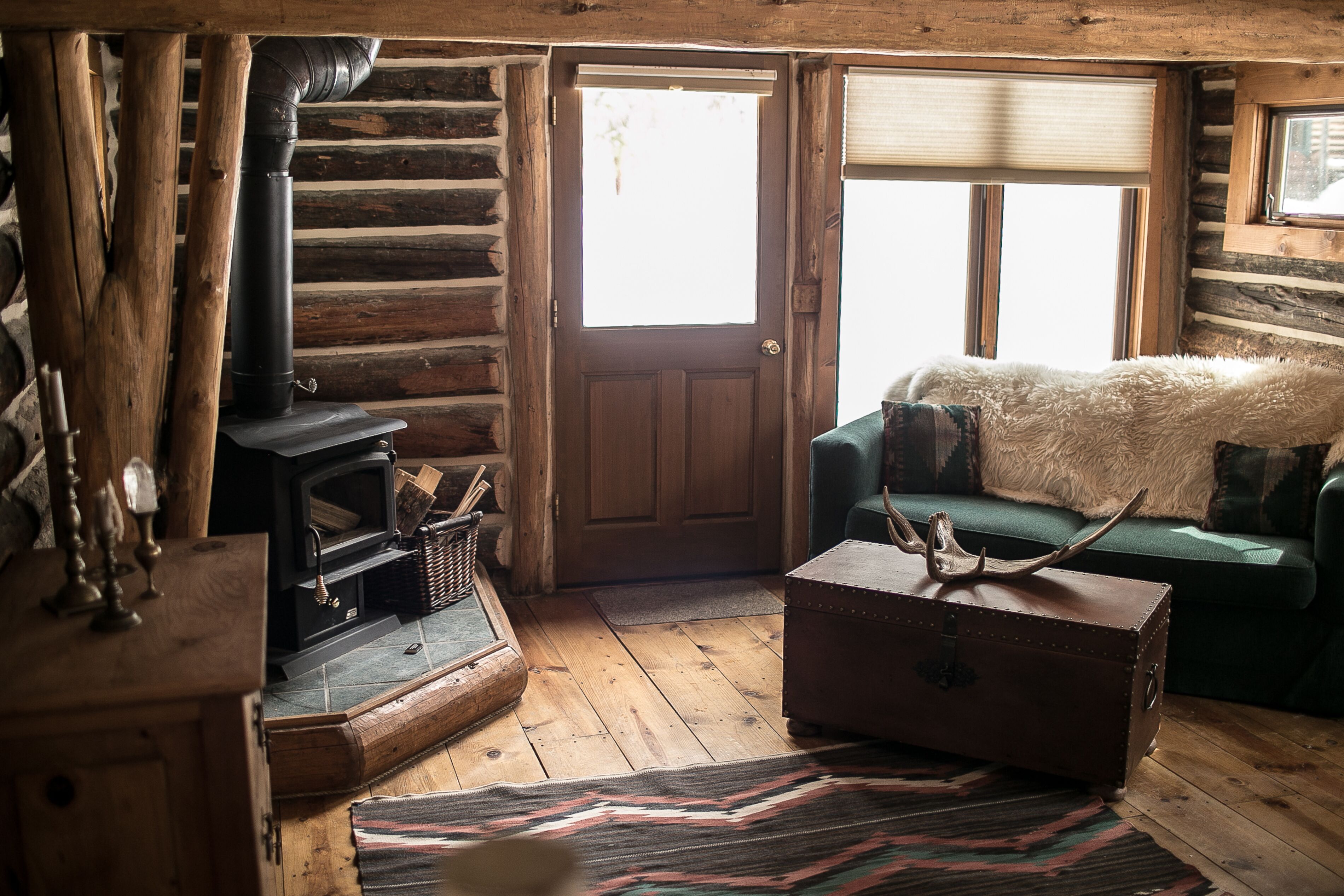 Ein rustikales Holzhütten-Interieur mit Holzofen, einem grünen Sofa mit Felldecke und einem alten Koffer.