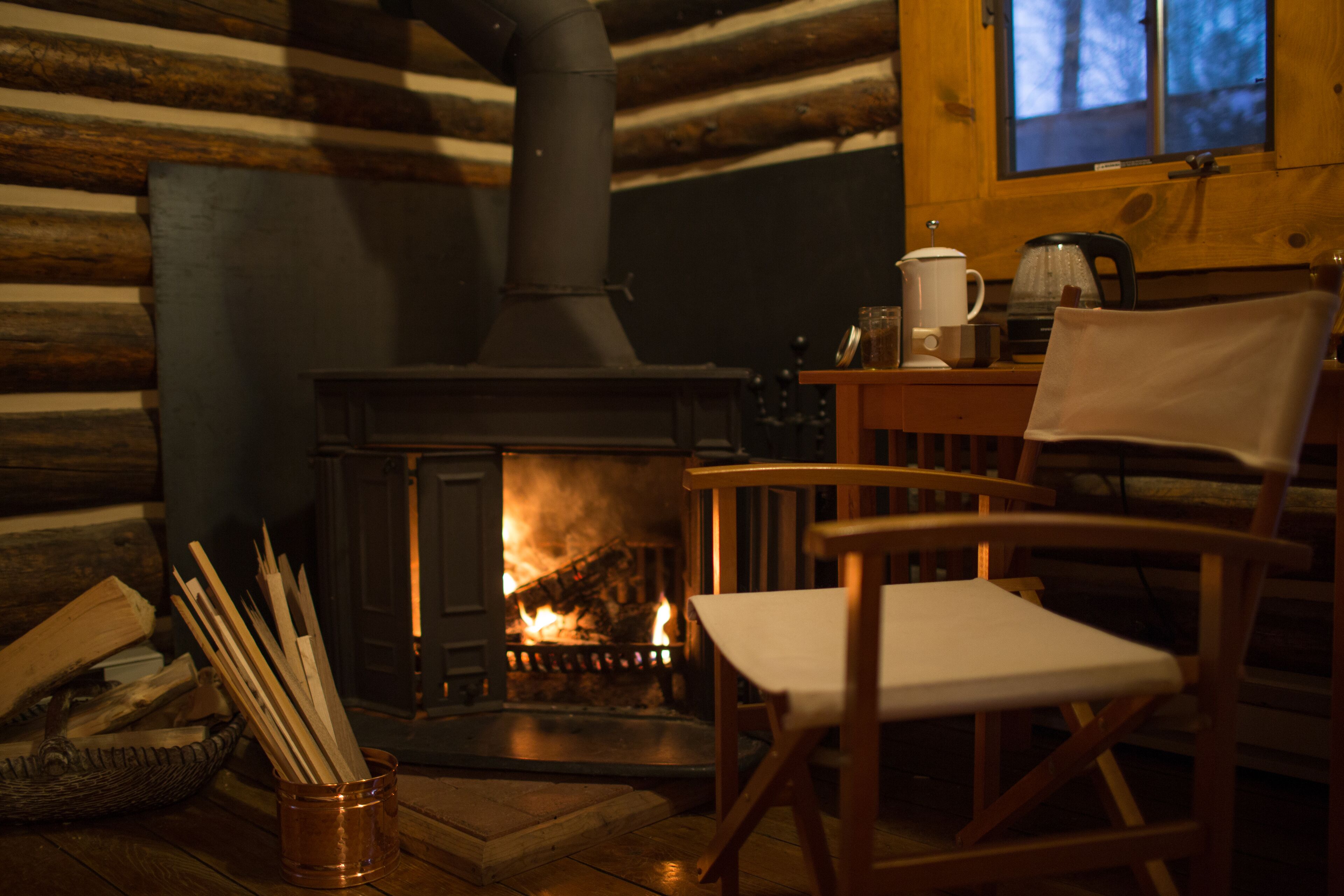 A cozy cabin interior with a wood-burning stove, a chair, and a window overlooking a forest.