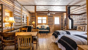 A log cabin room with a wooden dining table, chairs, a sofa, a bed with a black and white bedspread, a fireplace, and a window. - Beyul Retreat (Meredith)