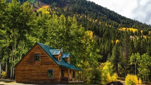 A log cabin with a green roof, surrounded by a forest and a mountain in the background. - Beyul Retreat (Meredith)