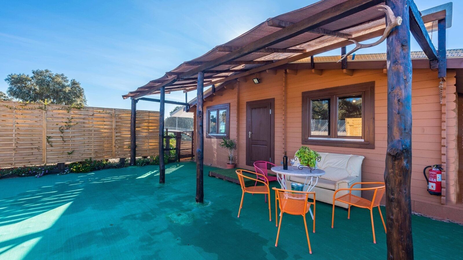 A covered outdoor seating area with a table and chairs, a fire extinguisher, and a wooden fence in the background.