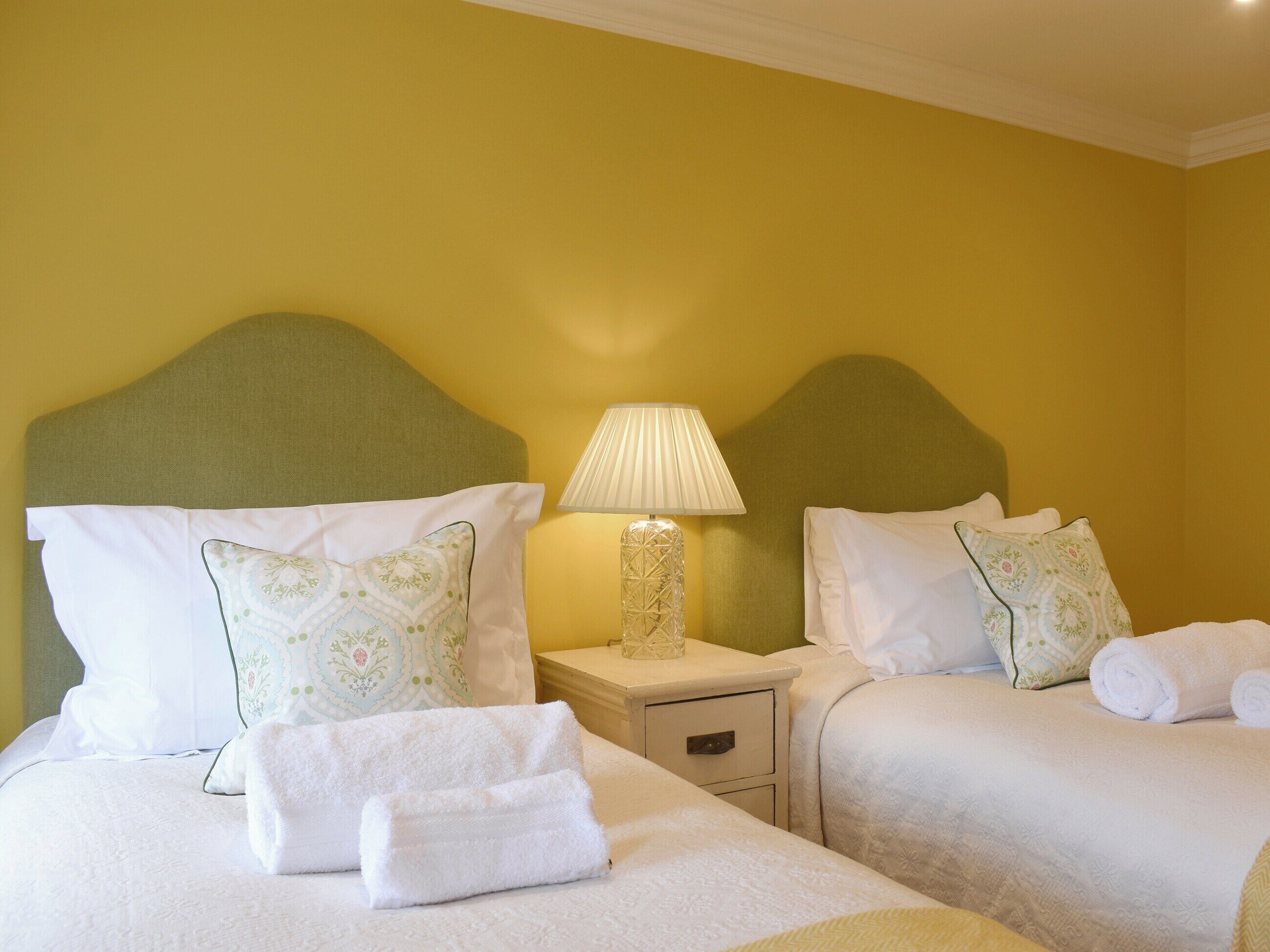 Une chambre d’hôtel avec deux lits, chacun agrémenté de draps blancs et d’oreillers assortis, une table de chevet avec une lampe, et un mur jaune.