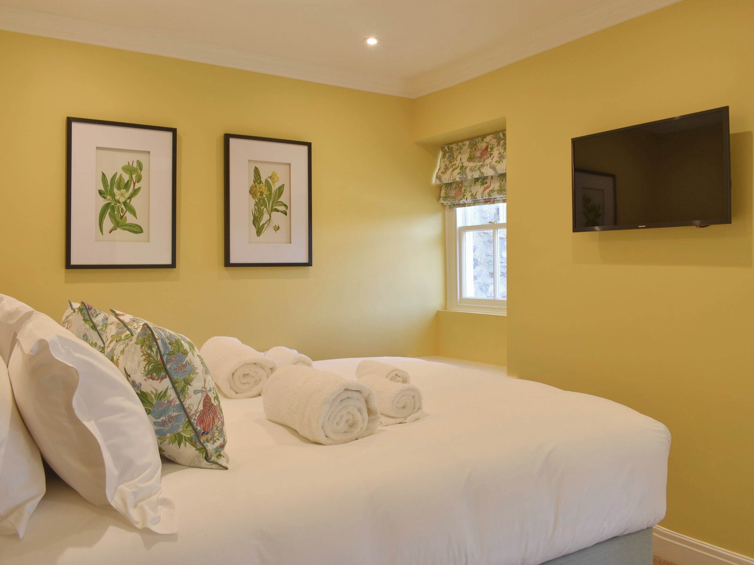 Una camera da letto con un letto grande, due stampe botaniche incorniciate alle pareti, un televisore montato sopra il letto e una finestra con tenda a fiori.