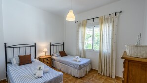 A room with two single beds, a wooden nightstand with a lamp, a window with curtains, and a wicker basket on a wooden dresser. - Casa Riu Tolox by Ruralidays (Málaga)
