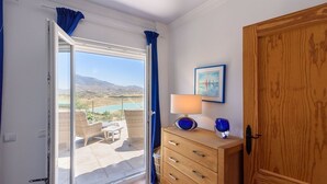 Ein Zimmer mit einer Glastür, die auf einen Balkon mit Blick auf Berge und einen See führt. Dort steht ein Holzkasten mit Schubladen und eine Lampe darauf.