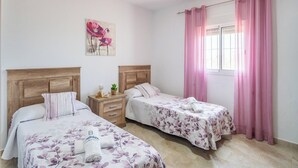 Een kamer met twee bedden, houten hoofdeinden, een klein nachtkastje met een lamp en een raam met roze gordijnen.