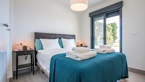 A neatly made bed with white and teal bedding, a bedside table with a lamp, and a view of greenery through a large window. - Capistrano Garden Nerja by Ruralidays (Málaga)