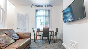 A living room with a brown leather sofa, a dining table with four chairs, a flat-screen TV mounted on the wall, and a window with blinds.