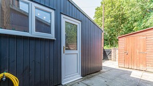 A dark-colored building with a white door and windows, a hose, and a wooden shed in the background.