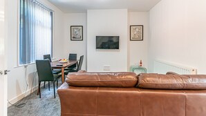 A living room with a brown leather sofa, a dining table with chairs, a TV mounted on the wall, and framed artwork on the walls.