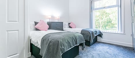 A bedroom with two beds, a window with blinds, a white door, and a carpeted floor.