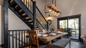 A dining area with a wooden table, chairs, and a bench. A staircase with black railings is visible to the left.