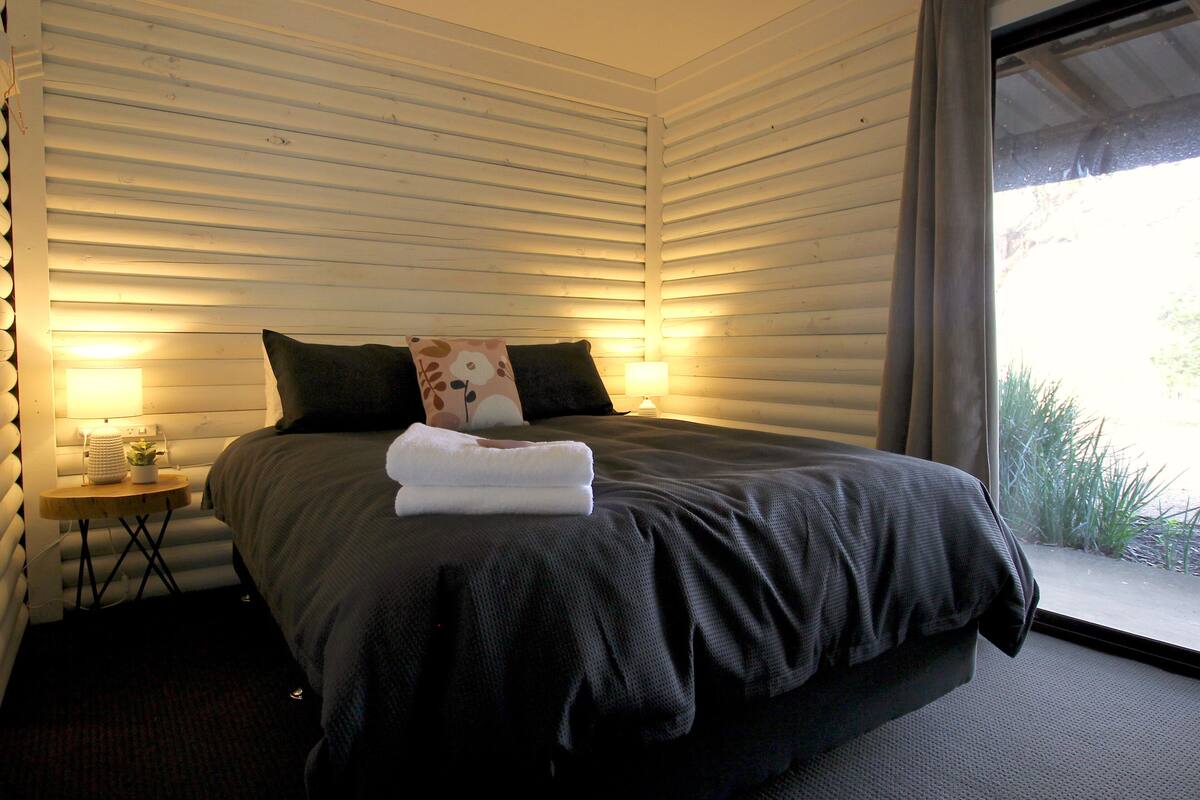 A bedroom with a bed, two bedside lamps, a small table with a plant, and a window with curtains.