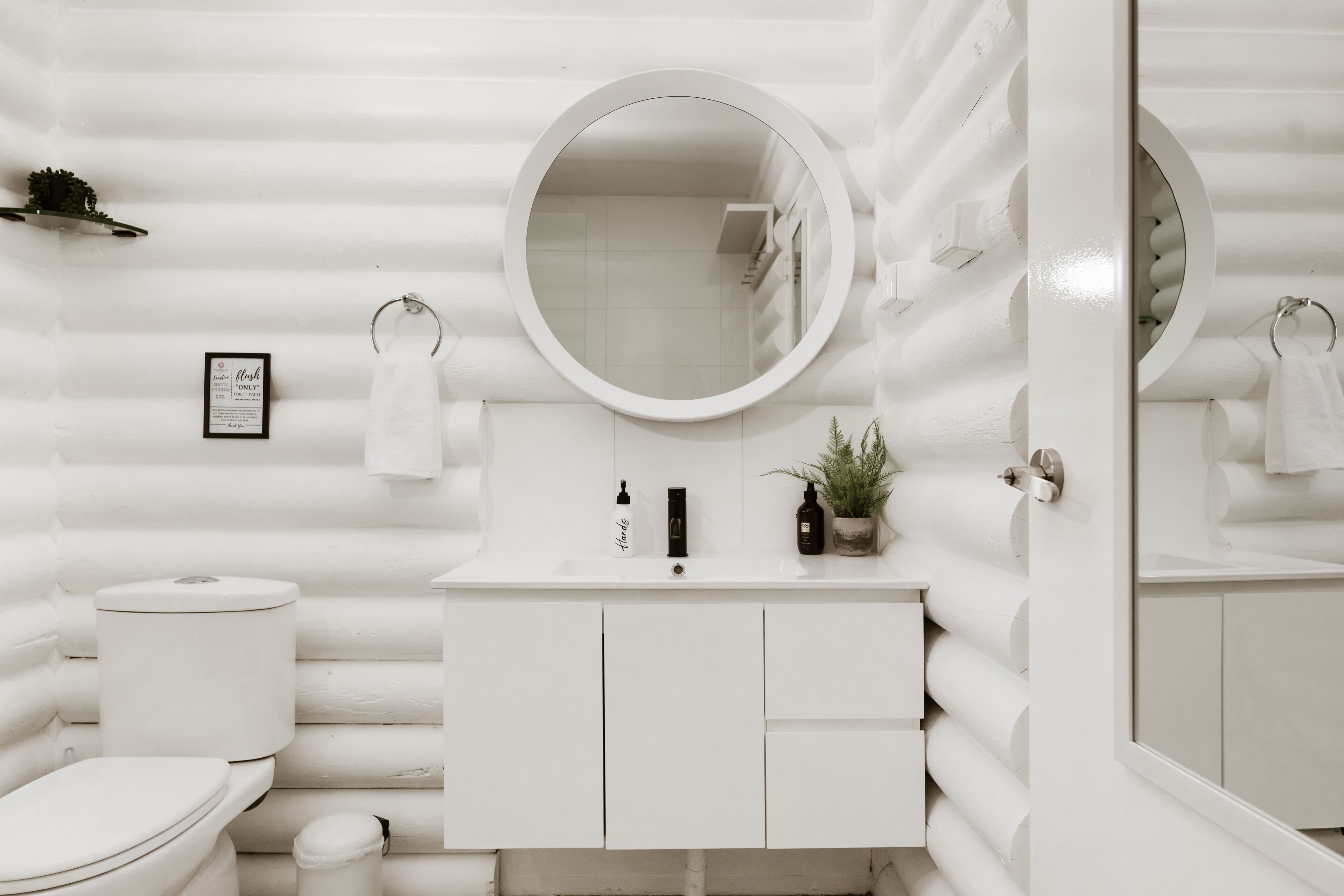 A white bathroom with a round mirror, a toilet, a sink with toiletries, and a small potted plant.