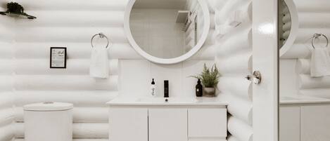 A white bathroom with a round mirror, a toilet, a sink with toiletries, and a small potted plant.