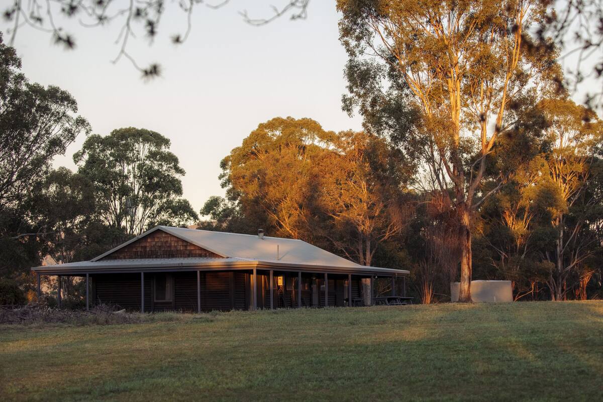 A rustic wooden house with a porch, surrounded by a grassy field and trees.