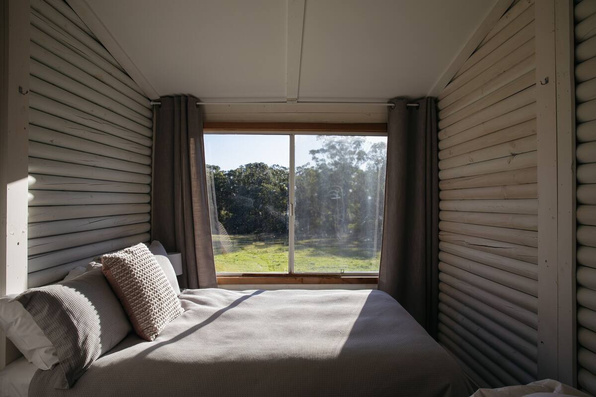 A bedroom with a large window, a bed with pillows, and a view of a green landscape.