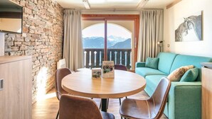 A cozy room with a stone wall, a round wooden table with chairs, a green sofa, and a view of mountains through a large window. - Sleeps 5! Mountain Escape in Valmorel | 1BR Apt (Doucy)