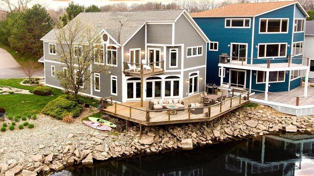 A two-story house with a deck overlooking a body of water, surrounded by a rocky shoreline.