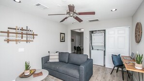 A modern living room with a gray sofa, a small round table, and a ceiling fan. There is a kitchen area with a refrigerator and a dining table with chairs. - 🏜Urban Desert Retreat for the Adventurous Traveler Snow Free-Stay with us! ☀️🐾 (Tempe)