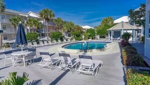 Un espace aménagé au bord de la piscine, avec des chaises longues et une table, entouré de palmiers et d’un bâtiment doté de balcons.
