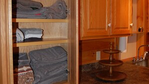 Un meuble en bois avec des serviettes pliées rangées sur des étagères, un coin cuisine avec un évier et un plan de travail, et une chaise.