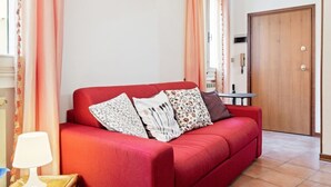 Un divano rosso con cuscini a fiori e a righe, un tavolino rotondo, una lampada e una porta in legno in una stanza con pareti bianche e pavimento piastrellato.