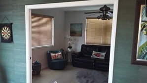 A living room with a black leather sofa, a blue armchair, and a grey rug. There are two windows with blinds, a ceiling fan, and a framed picture on the wall. - Paradise on the Canal with Ocean Access including Pool & Spa (Key Largo)