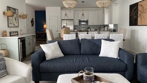 Un salon moderne avec un canapé bleu foncé, des coussins blancs à motifs, une table basse en bois et une cuisine en arrière-plan.