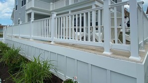 Une maison à deux étages, avec une façade blanche, un balcon doté de balustrades blanches et un jardin où poussent des plantes vertes et des fleurs roses.